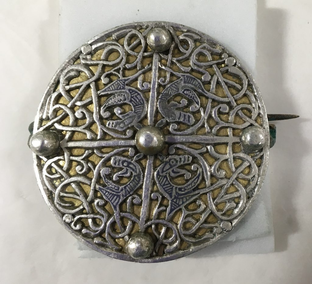 The almost fully conserved circular brooch viewed head-on against a white surface. All traces of green corrosion are gone, emphasising the silver interlace pattern and gold-coloured base.