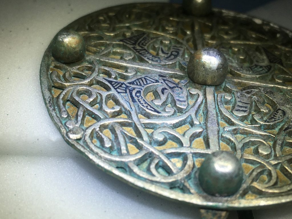 Closeup of the bottom half of the circular brooch, highlighting corrosion along the edges of the metal interlace decoration.