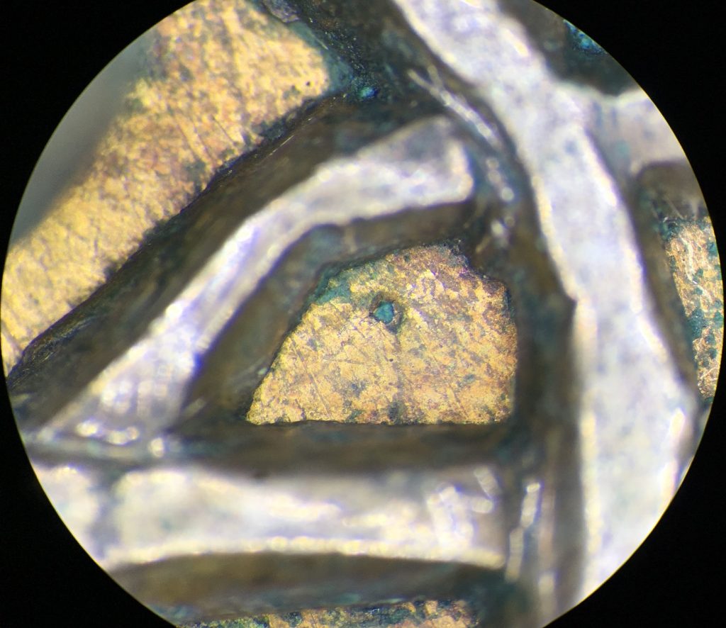 Extreme microscopic closeup of a triangular section of copper framed by silver lines. Tiny green dots of corrosion break through the copper in several places.