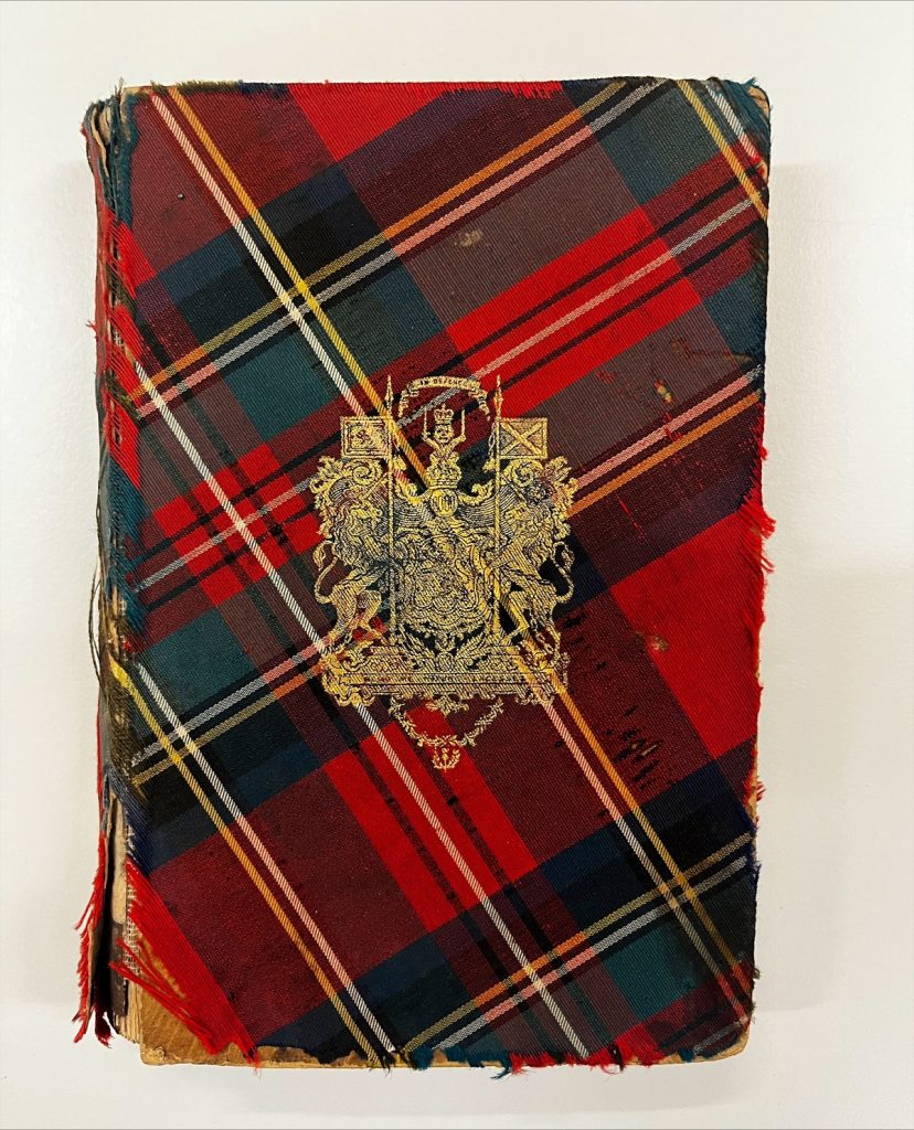 The cover of a book wrapped in a red and green tartan, with an intricate golden coat-of-arms in the centre. No title is displayed.