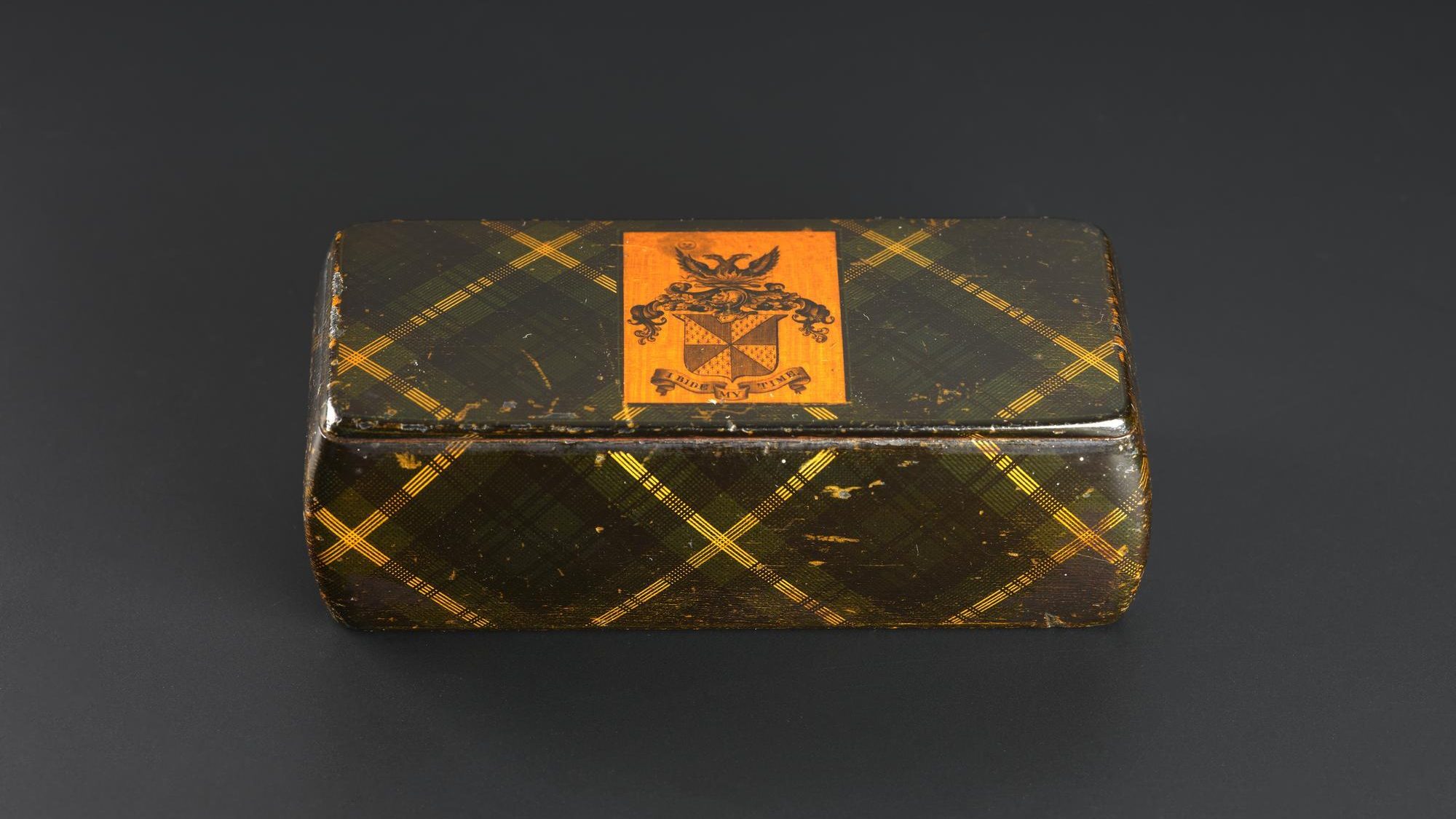 A small, rectangular snuffbox against a grey background. The snuffbox is covered in a green and yellow-striped tartan pattern, and has a yellow square with a black coat of arms in the centre of the lid.