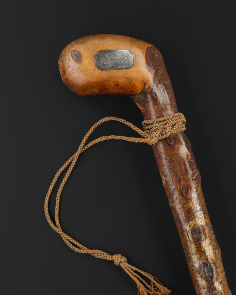Top half of a dark brown ridged wooden walking stick with a curved top. A metal plate on the handle of the stick reads “Sir Walter Scott Abbotsford”.