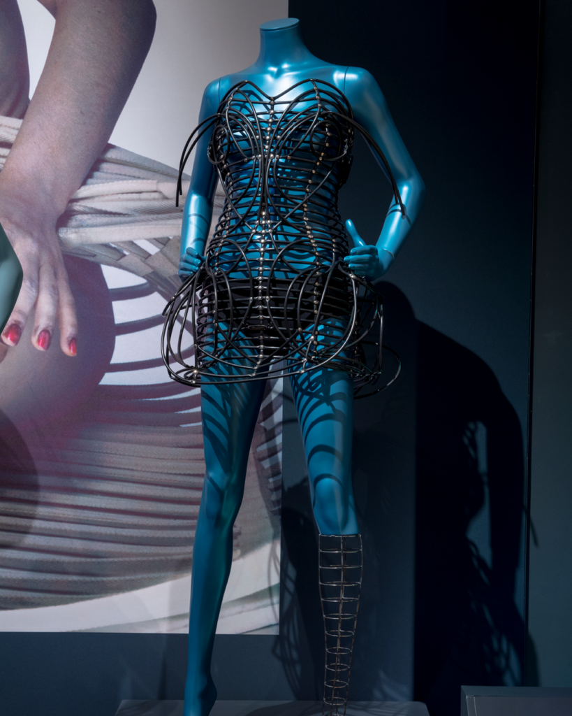 Black mini dress with heart neckline and short rounded skirt made of a plastic cage construction. It is modelled on a blue mannequin whose left leg below the knee is replaced by a black cage structure.