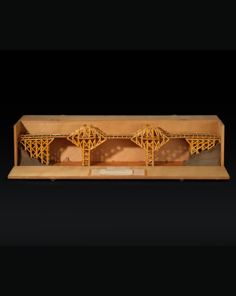 Model bridge made from light brown matchsticks with a grey train track down the middle of the bridge and housed in a brown wooden box.