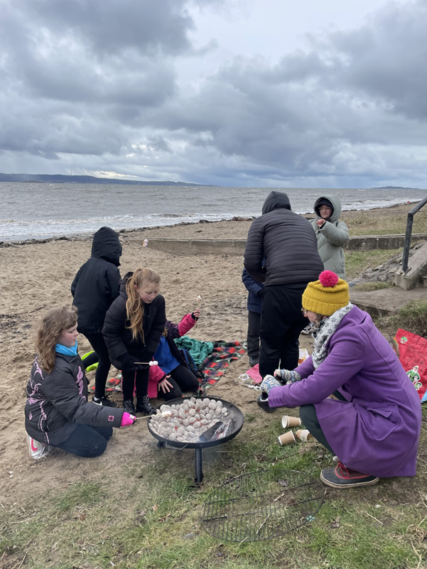 Six young people and two adults toast marshmallows on a beach.
