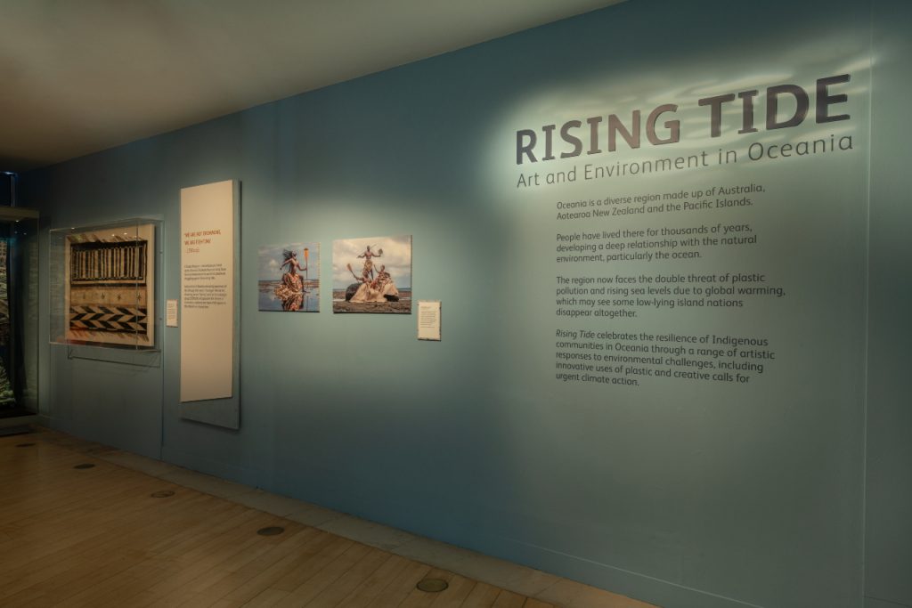 A view of the 'Rising Tide' exhibition, featuring wall text. 'Rising Tide Art and Environment in Oceania. Oceania is a diverse region made up of Australia, Aotearoa New Zealand and the Pacific islands. People have lived there for thousands of years, developing a deep relationship with the natural environment, particularly the ocean. The region now faces the double threat of plastic pollution and rising sea levels due to global warming, which may see some low-lying island nations disappear altogether. Rising Tide celebrates the resilience of Indigenous communities in Oceania through a range of artistic responses to environmental challenges, including innovative uses of plastic and creative calls for urgent climate action. 