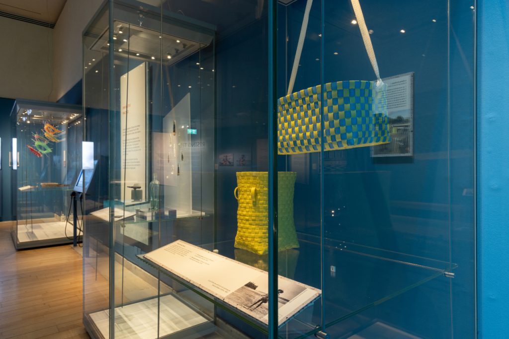 Cases in the 'Rising Tide' exhibition, including two brightly coloured baskets woven with yellow and yellow and blue plastic. 