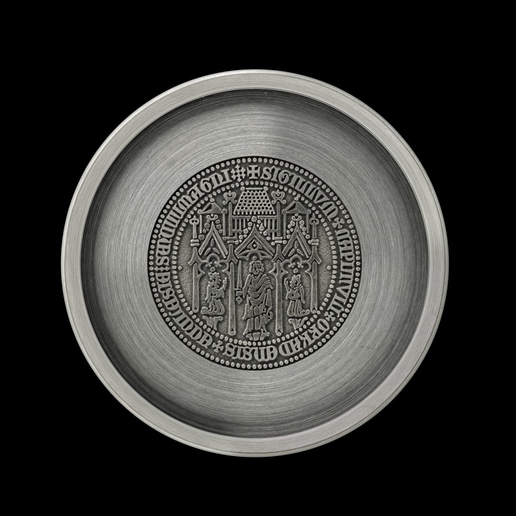 A circular brushed pewter coaster with a circular design in the centre showing three robed figures, monks, within a gothic arched architectural structure. With two figures kneeling in prayer on either side, the central figure wields a sword.