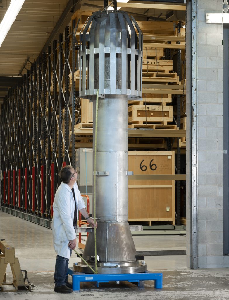 A large metal flare tip pictured in the museum collections centre. It's tall, cylindrical with a grilled 'cage' at the top. A person in a white coat is standing next to it, measuring it with a yellow measuring tape. For scale, the flare tip is over twice his height. 